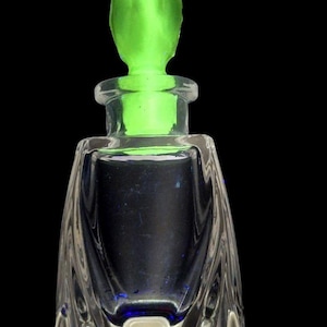 Puede incluir: Un frasco de perfume de vidrio transparente con un tapón verde. El frasco tiene una base cuadrada y un cuerpo acampanado y curvado. El tapón es de un verde vibrante, sobre un fondo negro.