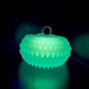 May include: A glowing green glass trinket box with a lid and a handle. The box has a textured, diamond-like pattern around the sides and lid. The handle is a small, circular ring. The background is dark.