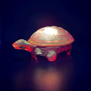 Puede incluir: Una figura de tortuga de vidrio translúcido iluminada desde el interior, que proyecta un resplandor rojo vibrante. El caparazón presenta un patrón geométrico detallado en relieve, mientras que la cabeza, las patas y la cola son lisas.