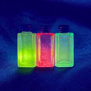 May include: Three rectangular bottles with black caps, each filled with a different colored liquid: green, red, and green. The bottles are illuminated, creating a glowing effect against a dark blue background.