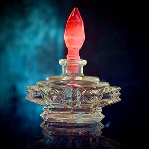 Könnte beinhalten: Eine klare Parfümflasche aus Glas mit einem roten, spitzen Verschluss. Die Flasche hat ein dekoratives, geometrisches Design und steht auf einer reflektierenden Oberfläche. Der Hintergrund ist dunkel mit einem blauen Farbverlauf.