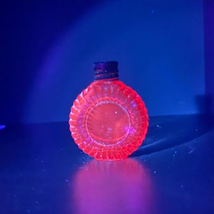 Puede incluir: Una pequeña botella de vidrio redonda y translúcida que brilla con un tono naranja neón vibrante bajo luz ultravioleta. La botella presenta un patrón exterior texturizado y acanalado a lo largo de su borde circular y una tapa oscura.