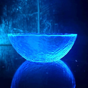 May include: A glowing blue glass bowl with a textured surface, reflecting on a dark surface. The bowl has a unique, organic shape and is illuminated with a bright blue light, creating a striking visual effect.