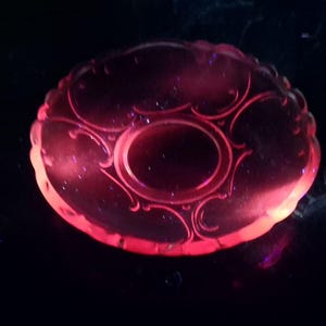 May include: A circular glass plate glowing with a vibrant pink hue under ultraviolet light. The plate features a central circular indentation surrounded by an embossed, symmetrical scrollwork pattern and a scalloped edge along the outer rim.