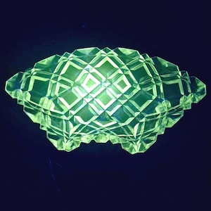 Può includere: Una ciotola di vetro verde brillante con un motivo geometrico a diamante. La ciotola ha una forma bassa e larga e si trova su uno sfondo scuro. Il vetro sembra essere vintage o antico.