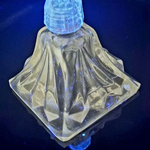 Puede incluir: Un frasco de perfume de vidrio transparente y facetado, con base cuadrada y cuerpo acampanado y drapeado. El frasco tiene un tapón cilíndrico y estriado. El vidrio refleja la luz azul.