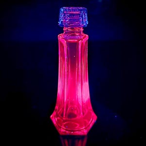 Könnte beinhalten: Eine hohe, transparente Glasflasche mit sechseckigem Boden und geriffeltem Körper. Sie ist mit einer rosa Flüssigkeit gefüllt und mit einem blauen, strukturierten Verschluss versehen. Das Bild vor dunklem Hintergrund, die Flasche ist beleuchtet, ca. 15 cm hoch.