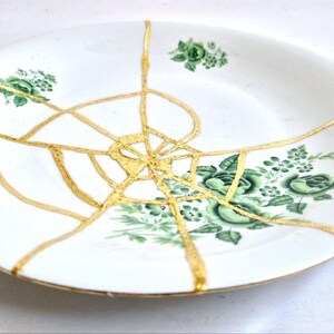 May include: White ceramic plate with green floral designs and gold-colored kintsugi repair. The plate features a round shape with a slightly raised edge. The gold lines highlight the repaired cracks, creating a unique decorative piece.