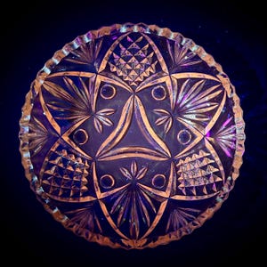 Könnte beinhalten: Eine klare Glasschale mit einem gewellten Rand und komplizierten geometrischen Mustern. Die Schale hat eine irisierende Oberfläche, die in Schattierungen von Lila, Rosa und Blau schimmert.