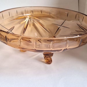 Könnte beinhalten: Bernsteinfarbene Servierschale aus Glas mit Sternmuster. Die runde Schale hat ein geometrisches Muster am Rand und wird von drei kleinen, abgerundeten Füßen getragen. Ideal für Obst oder Süßigkeiten.