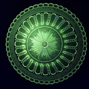 Könnte beinhalten: Ein kreisförmiger, grüner Glasteller mit einem komplizierten, geätzten Design. Der Teller weist ein zentrales Blumenmotiv auf, das von strahlenförmigen, bogenförmigen Mustern und einem Perlenrand umgeben ist. Das Glas hat einen leichten Glanz vor dem dunklen Hintergrund.