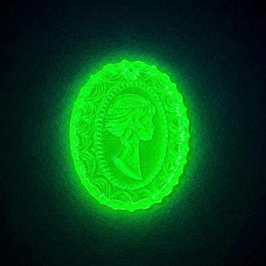 Könnte beinhalten: Ein ovaler Kamee-Anhänger, der in einem hellen Neongrün leuchtet. Das Stück zeigt ein detailliertes Profilrelief einer Person mit aufwendiger Frisur, umgeben von einem dekorativen, gewellten Rand mit geprägten floralen Mustern.