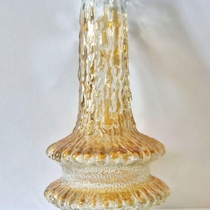 Pode incluir: Um vaso de vidro transparente com um exterior texturizado de cor âmbar. O vaso tem uma base larga, uma seção média bulbosa e um pescoço alto e fino. A parte superior do vaso tem uma borda texturizada e irregular.