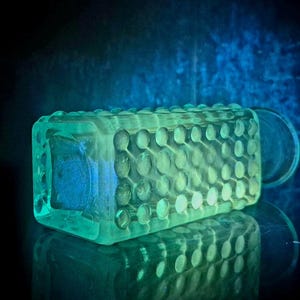 Op de afbeelding: Een rechthoekige glazen fles met een hobnail-patroon met verhoogde ronde bobbels over het oppervlak. Het glas straalt een heldere, lichtgevende neongroene gloed uit, die het getextureerde rasterontwerp tegen een donkere achtergrond benadrukt.
