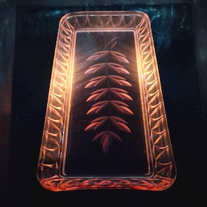 May include: A rectangular pink glass tray featuring a central etched leaf pattern and a decorative scalloped border along the rim. The translucent glass has a smooth interior surface and a textured, patterned edge that catches the light.