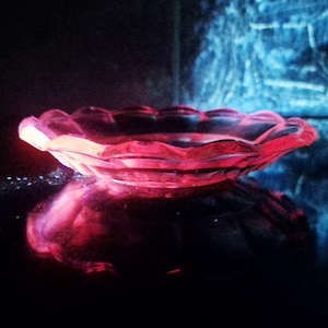 May include: A translucent pink glass dish with a scalloped edge and a shallow, circular basin. The glass glows with a vibrant pink hue, reflecting light across its surface, creating a distinct visual contrast against the dark surroundings.