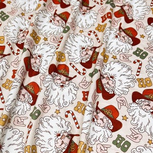 Puede incluir: Tela color crema con un estampado repetido de Papá Noel con sombrero de vaquero, bastones de caramelo, estrellas y la palabra "HO". El sombrero de Papá Noel y la palabra "HO" son rojos y dorados.