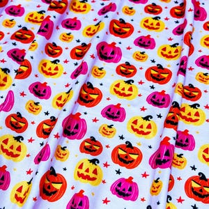 Puede incluir: Tela blanca con un patrón repetido de calabazas de Halloween de varios tamaños y colores. Las calabazas son naranjas, amarillas y rosas, con rasgos faciales negros. Pequeñas estrellas rojas y negras están dispersas.