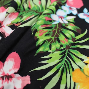 Puede incluir: Primer plano de una tela negra con un vibrante estampado floral. El diseño presenta grandes flores rosas, amarillas y blancas, junto con hojas verdes. La tela probablemente se utiliza para ropa o decoración del hogar.