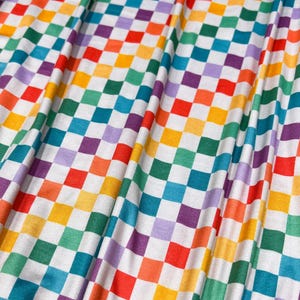 Puede incluir: Primer plano de una tela con un patrón de tablero de ajedrez arcoíris repetido. Los cuadrados son rojos, naranjas, amarillos, verdes, azules y morados, sobre un fondo blanco. La tela está drapeada en pliegues suaves.