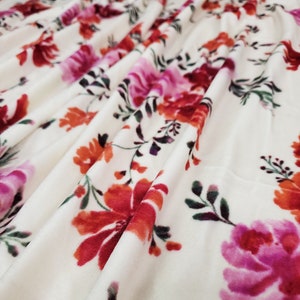 May include: White fabric with a floral print featuring pink, red, and orange flowers. The fabric has a soft, velvety texture.