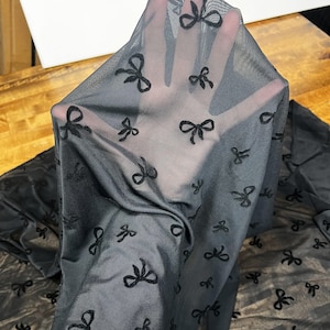 以下が含まれることがあります： 小さな刺繍リボンの繰り返し模様が施された黒のシアー生地。生地は持ち上げられ、光が通り、繊細なデザインを際立たせています。リボンは素材全体に均等に配置されています。