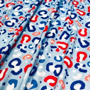 Puede incluir: Tela azul claro con un estampado de leopardo repetido en rojo, blanco y azul. La tela está doblada, mostrando la textura y el diseño del material. El patrón es una versión moderna de un estampado animal clásico.