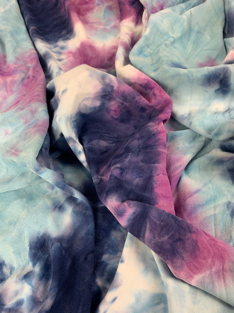 May include: A close-up of a tie-dye fabric in shades of blue, purple, and white. The fabric is soft and has a crinkled texture.
