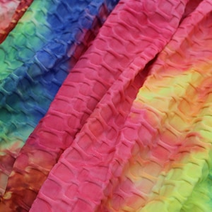 Op de afbeelding: Close-up van een kleurrijke tie-dye stof met een textuur, gestikt patroon. De stof is een mix van roze, blauw, geel en groen.