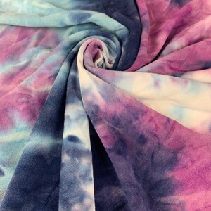 May include: A close-up of a tie-dye fabric in shades of blue, purple, and white. The fabric is soft and has a slightly textured surface.
