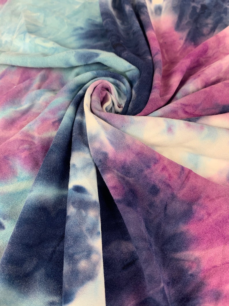 May include: A close-up of a tie-dye fabric in shades of blue, purple, and white. The fabric is soft and has a slightly textured surface.