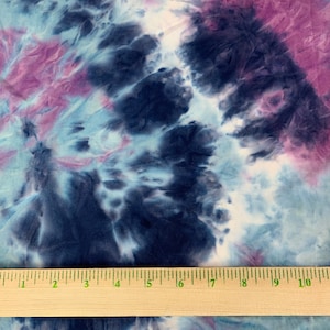 May include: A close-up of a tie-dye fabric with shades of blue, purple, and white. A wooden ruler is placed on the fabric to show the scale.