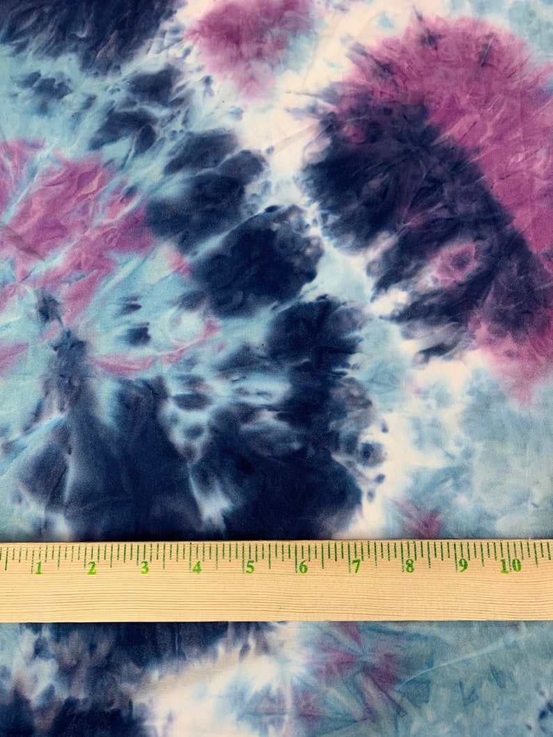 May include: A close-up of a tie-dye fabric with shades of blue, purple, and white. A wooden ruler is placed on the fabric to show the scale.
