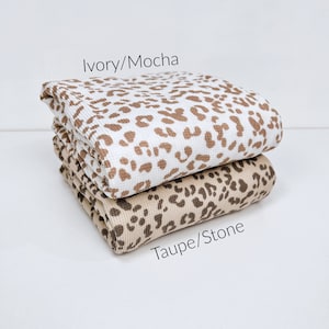 May include: Two folded pieces of fabric with a leopard print pattern. The top piece is ivory with brown spots and labelled "Ivory/Mocha". The bottom piece is taupe with brown spots and labelled "Taupe/Stone".