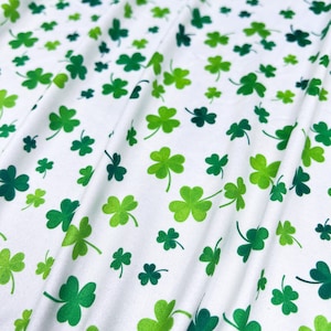 Puede incluir: Tela blanca con un patrón repetido de tréboles verdes en varias tonalidades. Los tréboles están dispersos por la tela, creando una apariencia texturizada. La tela está doblada, mostrando el flujo del patrón.