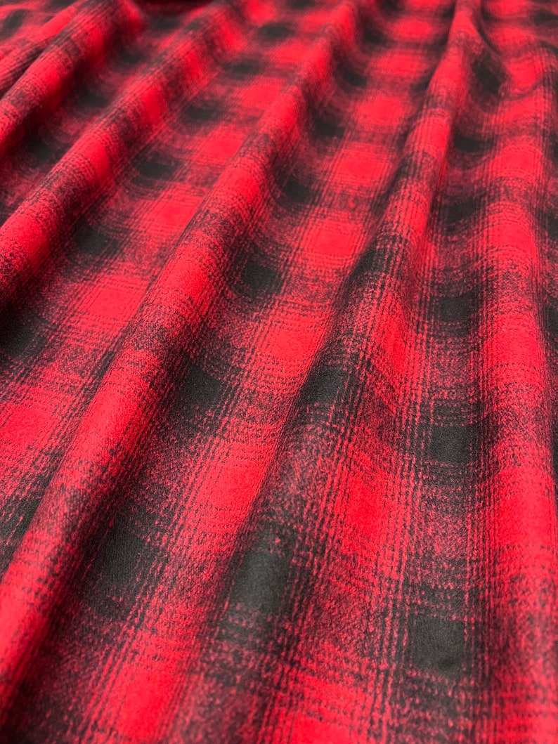 K&ouml;nnte beinhalten: Roter und schwarzer karierter Flanellstoff mit weicher, flauschiger Textur. Der Stoff ist gefaltet und drapiert, wodurch Muster und Textur sichtbar werden.