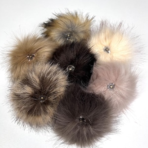 Puede incluir: Surtido de siete pompones de piel sintética en varios colores, incluyendo marrón, negro, beige y gris. Cada pompon tiene un pequeño botón en el centro.