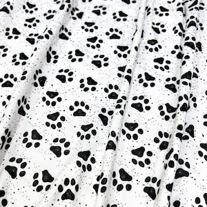 May include: A white fabric featuring a repeating pattern of black paw prints and small black speckles. The material is draped to show folds and texture, highlighting the consistent distribution of the animal-themed graphic print across the surface.