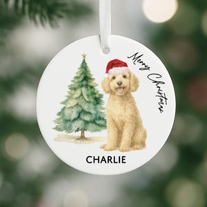 May include: A round, white Christmas ornament featuring a watercolor illustration of a golden doodle wearing a Santa hat, a Christmas tree, and the text "Merry Christmas." The dog's name, "Charlie," is printed below. A white ribbon is attached for hanging.