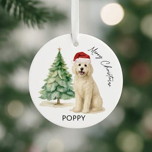 May include: Round Christmas ornament featuring a watercolor illustration of a dog wearing a Santa hat next to a Christmas tree. The ornament has the text "Merry Christmas" and the dog's name, "POPPY". A white ribbon is attached for hanging.