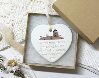 May include: White ceramic heart-shaped ornament with a brown house and trees illustration. The ornament has a string for hanging and the text "May your troubles be less your blessings be more and nothing but happiness come through your door".