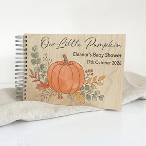 May include: A spiral-bound wooden guest book featuring a printed illustration of an orange pumpkin surrounded by autumn leaves and berries. The text reads Our Little Pumpkin, Eleanor's Baby Shower, and 17th October 2026 in black lettering.