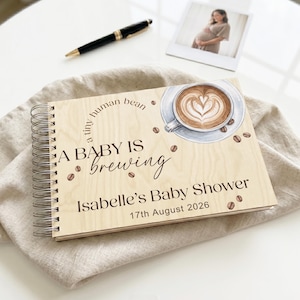 May include: A spiral-bound wooden guest book featuring a printed illustration of a coffee cup with latte art, scattered coffee beans, and the text: a tiny human bean, A BABY IS brewing, Isabelle's Baby Shower, and 17th August 2026.