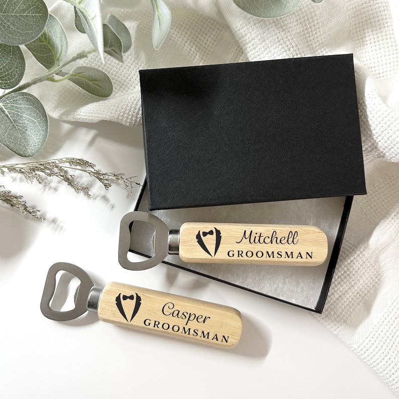 May include: Two wooden bottle openers with a tuxedo design and the words "Groomsman" and the names "Mitchell" and "Casper" engraved on them.