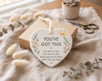 May include: A white heart-shaped ceramic plaque with a cream ribbon hanger. The text reads: YOU'VE GOT THIS, One day at a time, One step at a time, Keep going you're doing better than you think. It features floral illustrations and a small sun.
