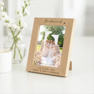 May include: A wooden picture frame with the word "Bridesmaid" in script. The frame displays a wedding photo. The date "31st October 2026" and names are printed at the bottom. A vase of white flowers is in the background. The frame is made of wood.