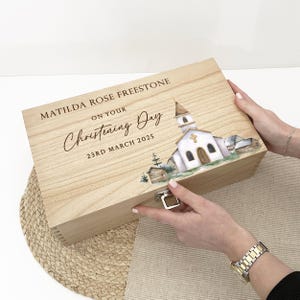May include: A light-coloured wooden keepsake box with a hinged lid. The lid features the text "MATILDA ROSE FREESTONE ON YOUR Christening Day 23RD MARCH 2025" and a watercolour illustration of a church. A silver clasp secures the box.