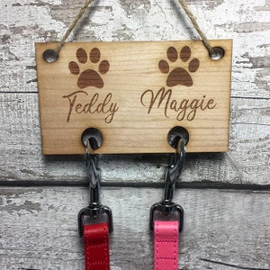 Pu&ograve; includere: Porta-guinzagli per cani in legno con due ganci e disegni di impronte di zampe. Il porta-guinzagli &egrave; inciso con i nomi "Teddy" e "Maggie".