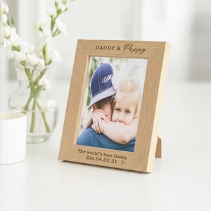 May include: A light-colored wooden picture frame with the words "DADDY & Pappy" engraved at the top. The frame holds a photo of a person hugging a child. The text "The world's best Daddy Est.04.03.21" is at the bottom.
