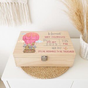 May include: A rectangular wooden keepsake box with a metal latch. The lid features a pink hot air balloon with animal illustrations, and printed text: Harriet, May Fieldhouse, Born on 6th July 2025, 7:25 P.M., 6lb 2oz, Special Memories To Be Treasured.